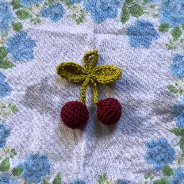 A crocheted cherry charm with a yellow-green leafy stem and two red cherries, placed on a white fabric cloth with a blue rose pattern.