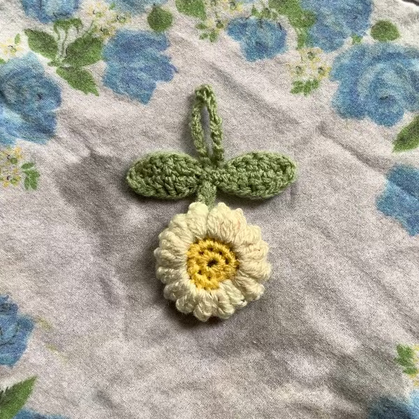 A handmade crocheted flower charm with green leaves and a cream-white daisy center lies on top of a square floral fabric with blue roses and green leaves.
