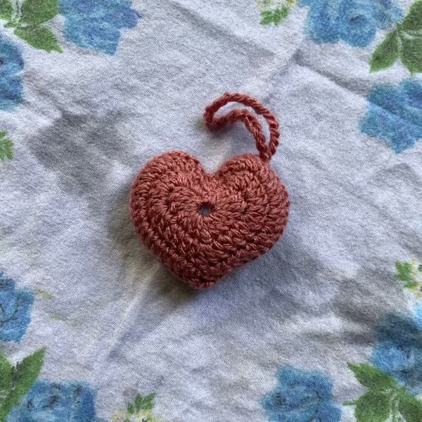 A small crocheted heart-shaped ornament in a terracotta rust color, resting on a white fabric background with a blue and green floral print.