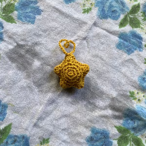A small crocheted star-shaped ornament in mustard yellow with a hanging loop, placed on a light gray fabric surface with a blue and green floral print.